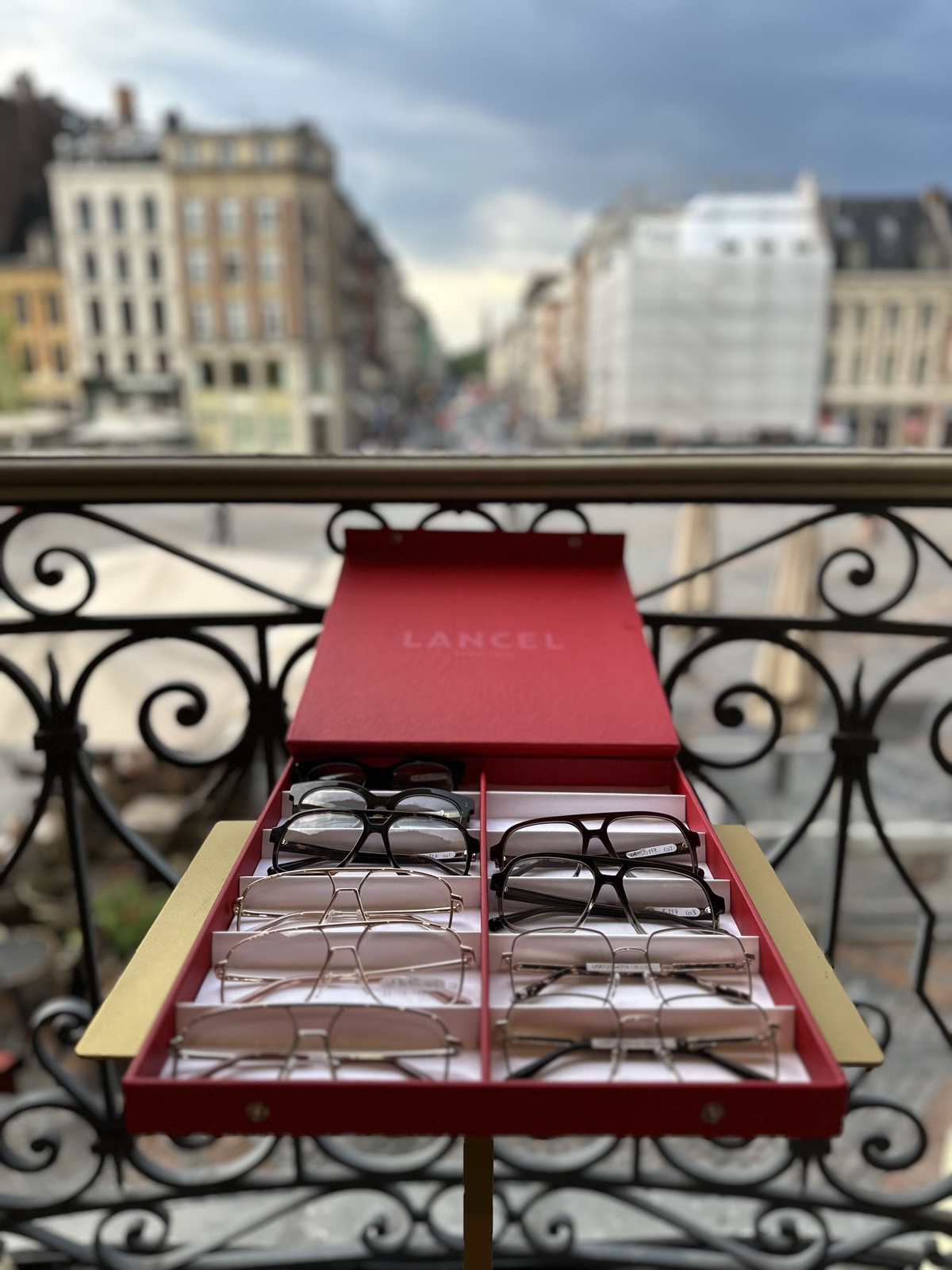 Un présentoir Lancel ouvert sur un balcon en fer forgé, ses montures alignées sur du velours rouge, une rue de Lille en contrebas