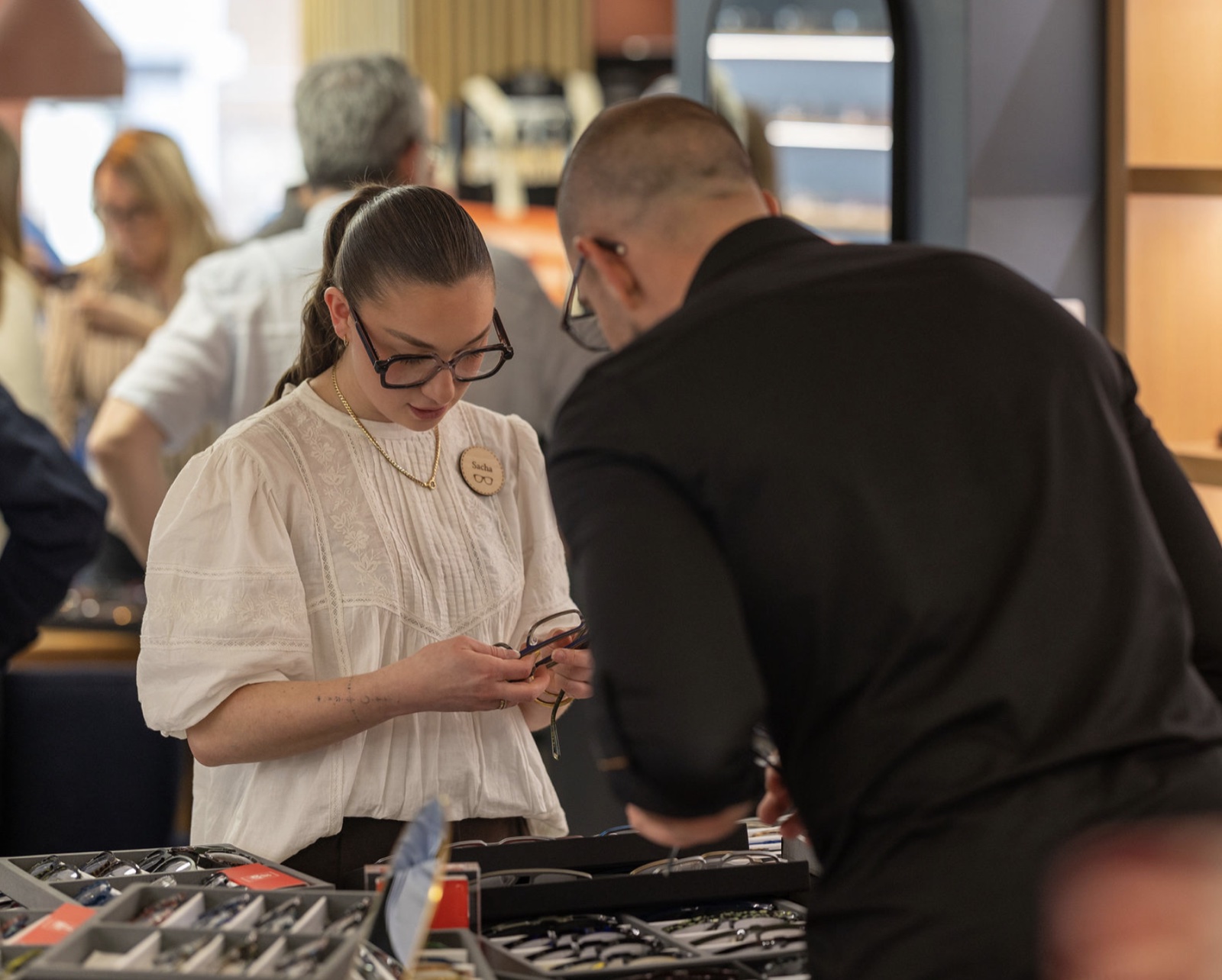 Une opticienne de l'équipe examine une monture avec un client, au-dessus des plateaux de collections posés sur le comptoir