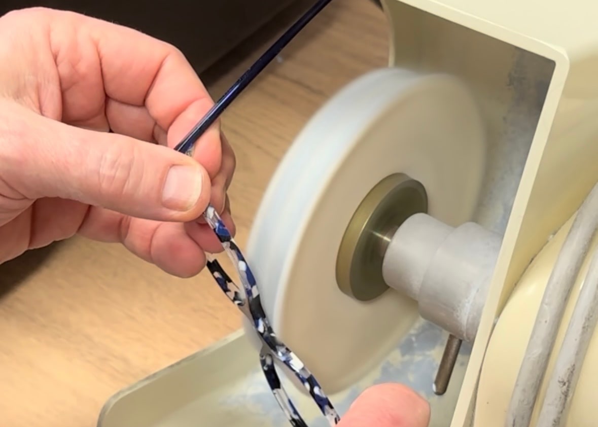 Deux mains présentent une monture en acétate bleu à la meule d’une machine d’atelier, chez Gravelines Optique