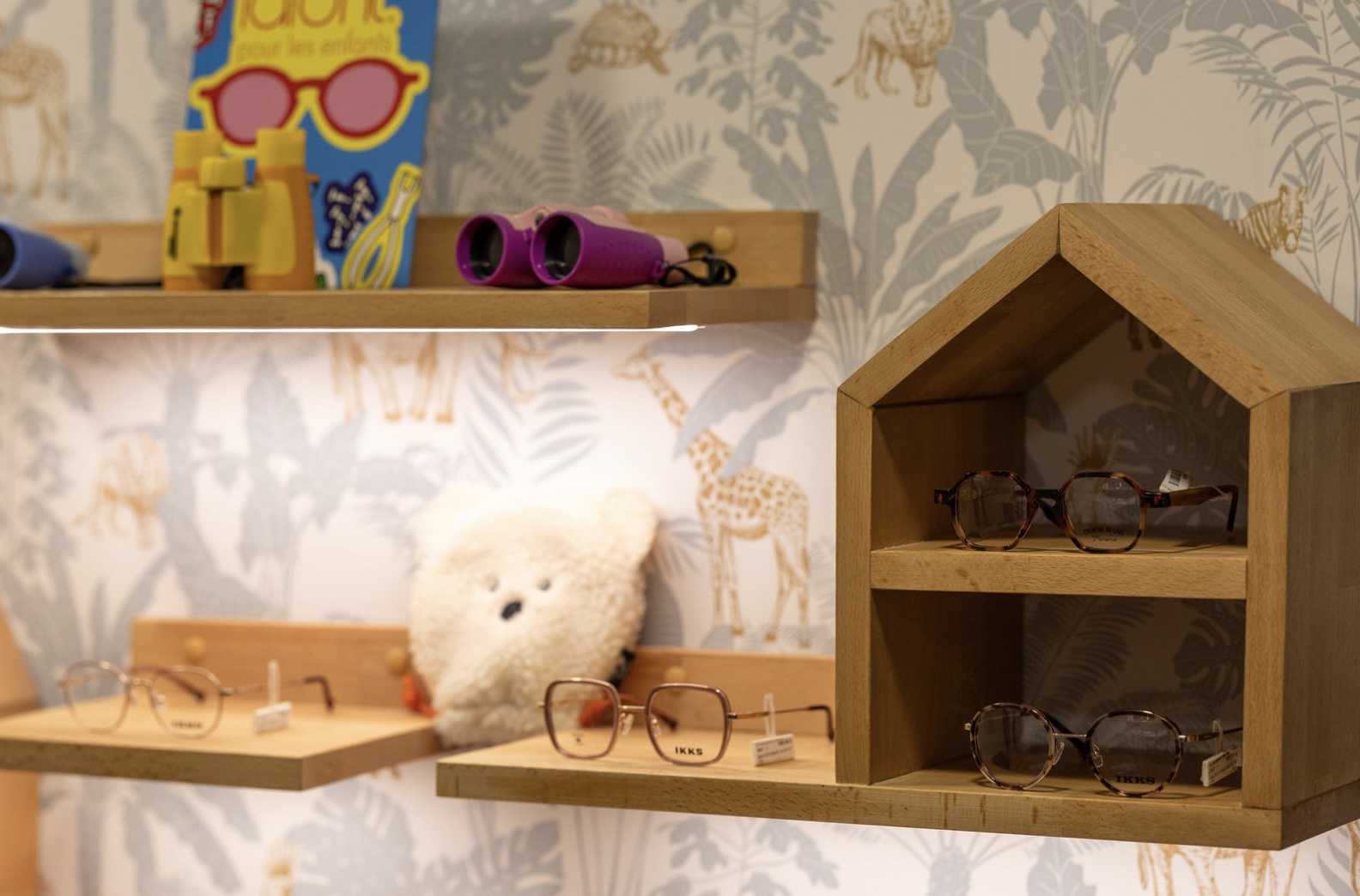 Le coin enfant de la boutique : montures IKKS sur un présentoir en forme de maison, papier peint jungle, jumelles jouet et peluche