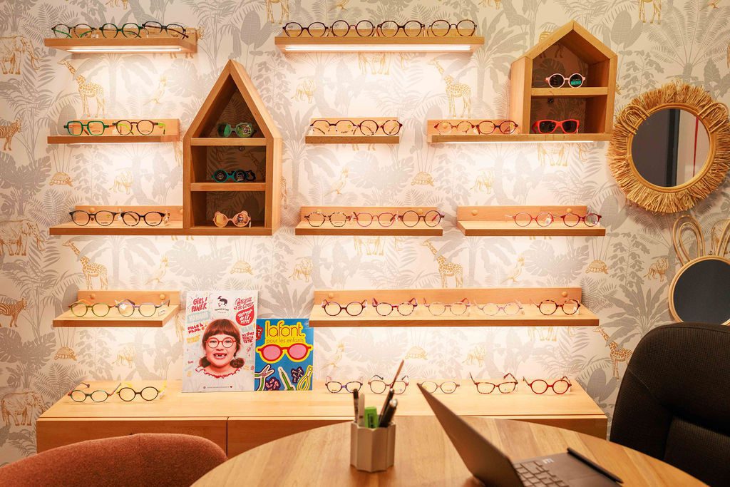 Le coin enfant de la boutique : des montures alignées sur des étagères de bois clair, des présentoirs en forme de maison et un papier peint à animaux