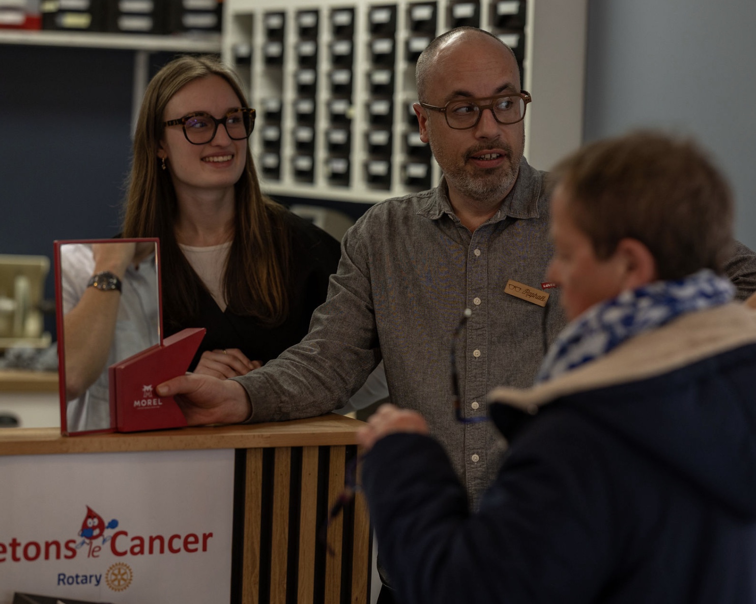 Raphaël remet une boîte Morel à un client au comptoir, une collègue à ses côtés, chez Gravelines Optique